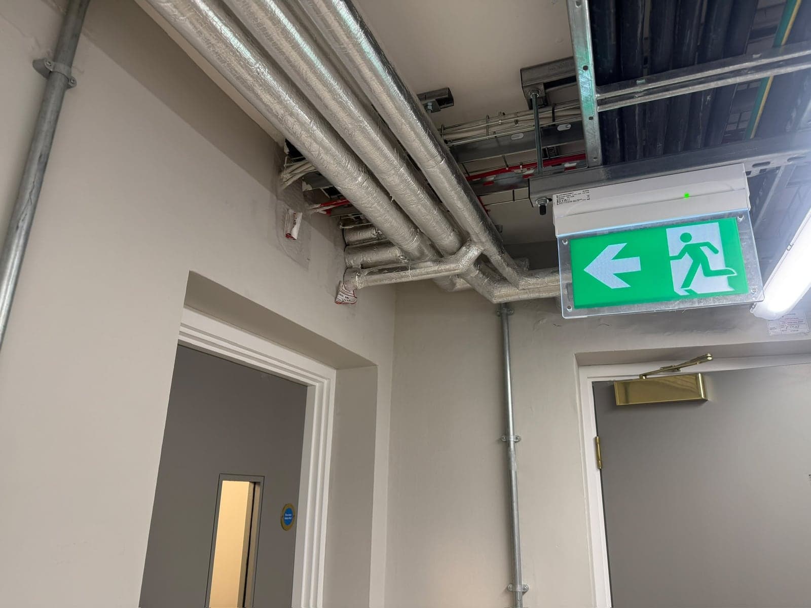 Emergency exit sign fitted under steel cable tray and conduit in a corridor